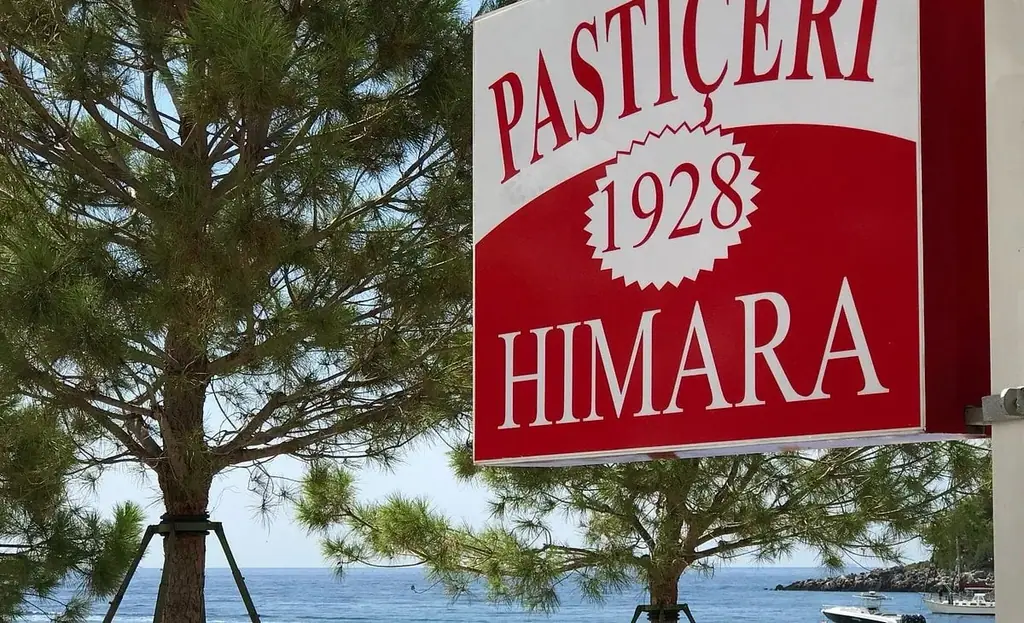 The painted board of Kafe Pastiçeri Himara 1928 on its post above the water at Spile, pines in front of it and boats moored beyond
