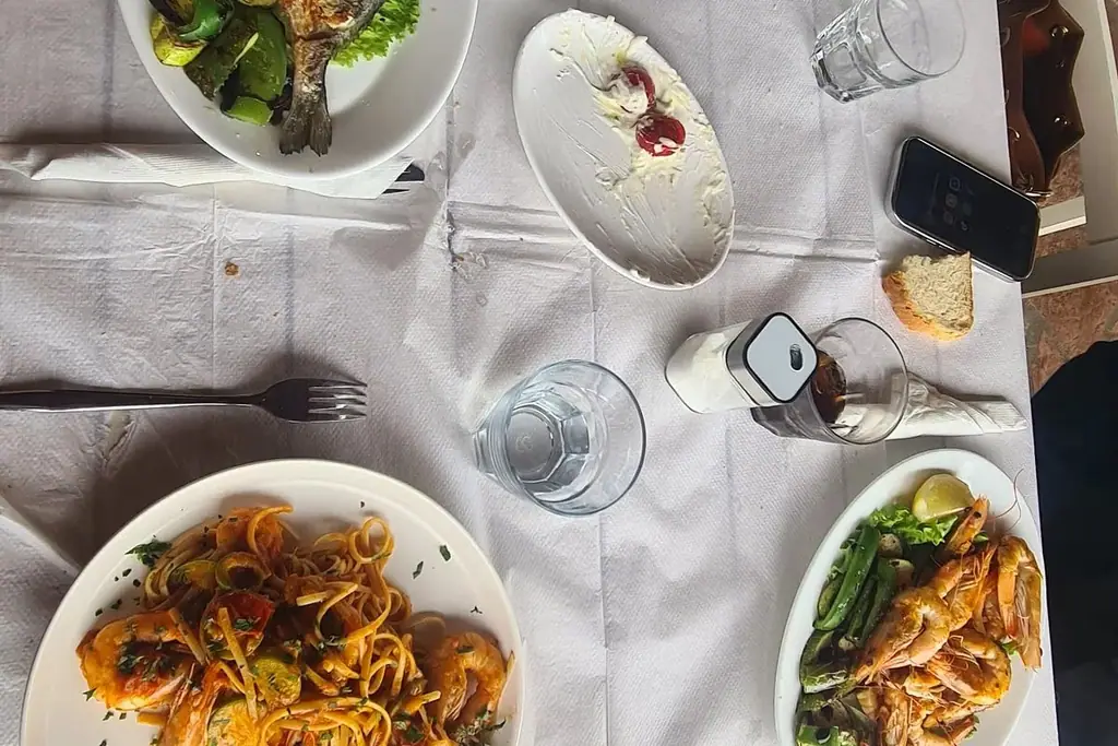 A table mid-meal seen from directly above: a grilled fish over grilled vegetables, linguine with prawns in tomato sauce, a plate of grilled prawns with lemon and a part-eaten plate of whipped cheese, on paper over the cloth