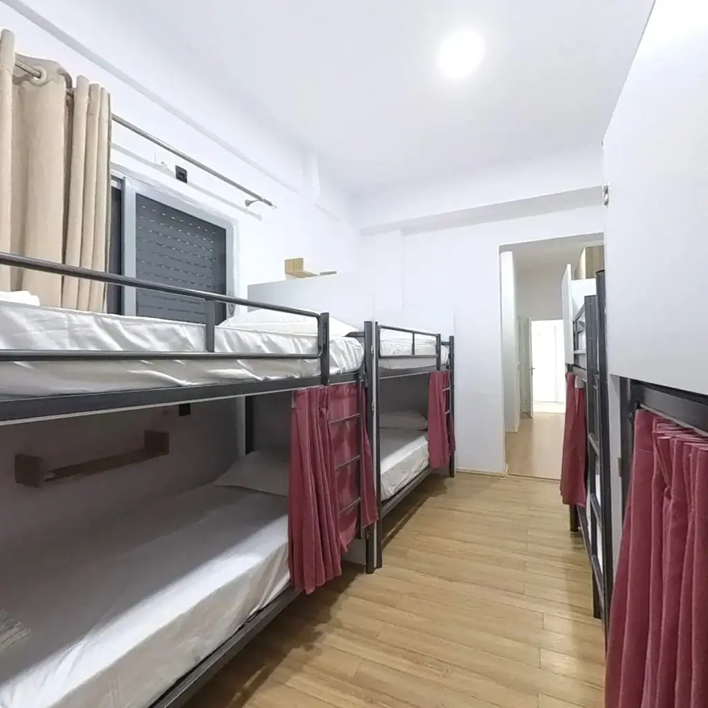A dormitory with steel bunk beds down both walls, dark red privacy curtains and a pale wooden floor