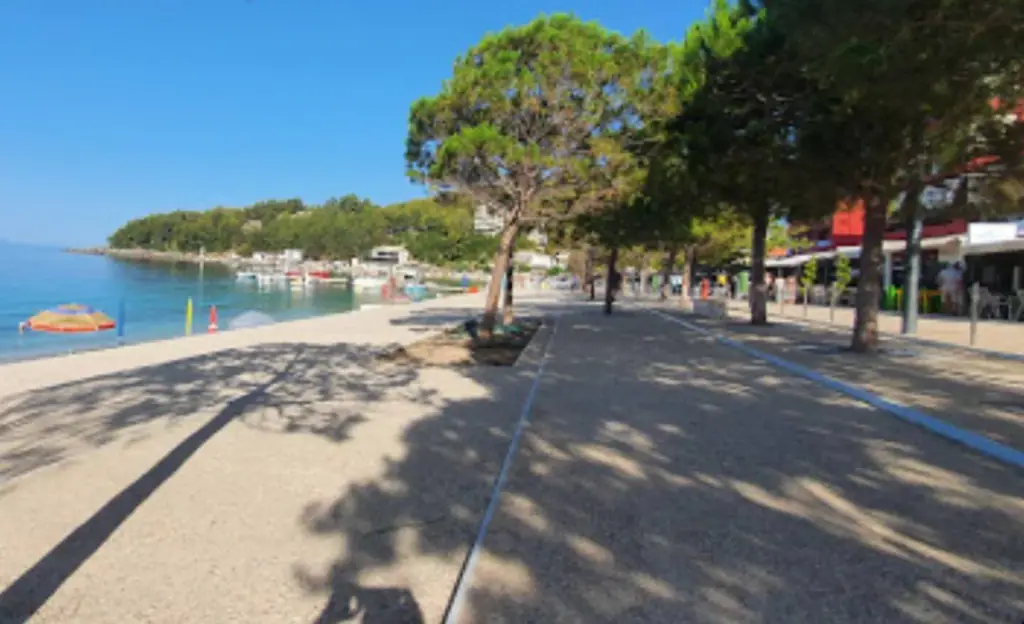Himarë's seafront promenade with umbrella pines, paving, the sea on one side and cafés under awnings on the other