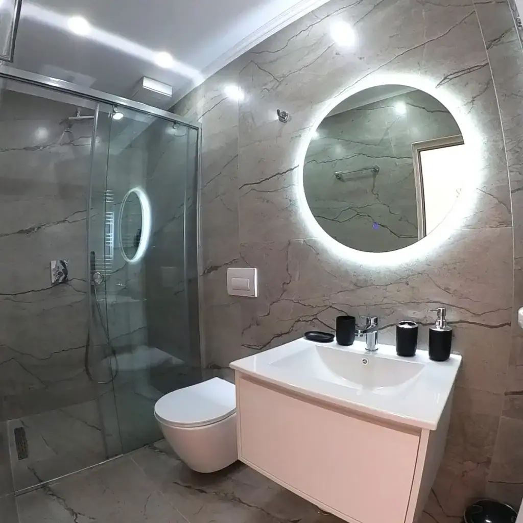 A grey marble bathroom with a glass walk-in shower, a wall-hung basin and a large round backlit mirror