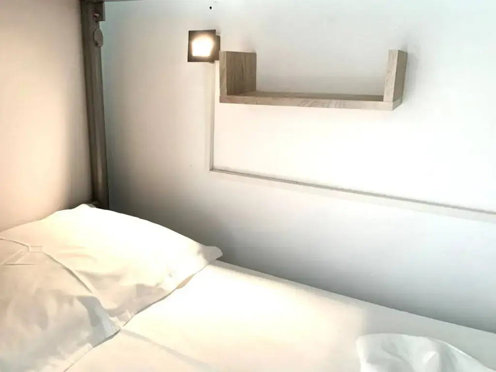 The inside of a lower bunk: white bedding, a small wall-mounted reading lamp lit, and a wooden shelf above the pillow