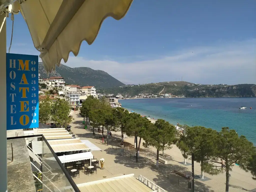 The blue Hostel Mateo sign in the foreground, with Himarë's promenade, umbrella pines, Spile Beach and the turquoise bay behind it