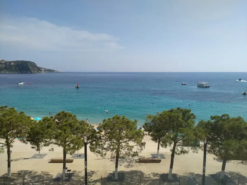 Spile Beach seen from above: umbrella pines along the promenade, pale sand, turquoise shallows, moored boats and the headland