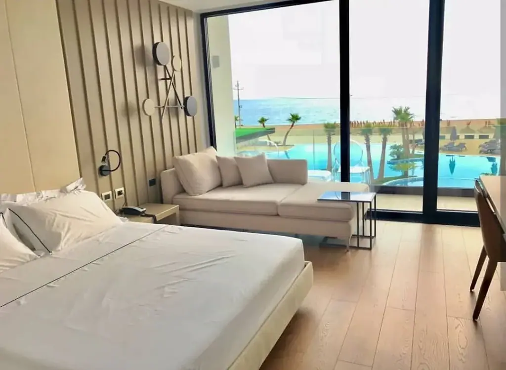 A guest room at Miamar with a king bed, a cream sofa and a full-height window onto the pool and the Ionian Sea