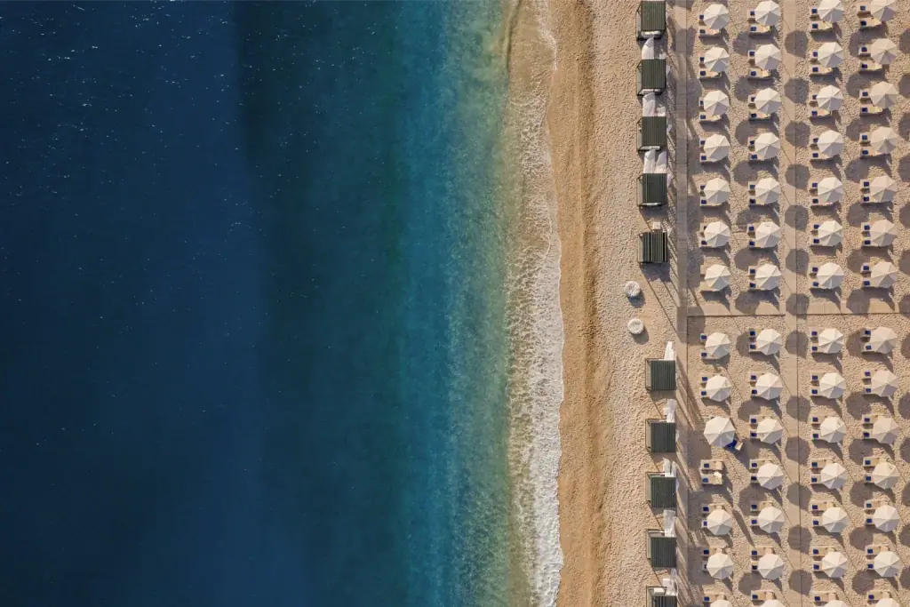 Livadhi beach from directly overhead — ruled rows of white parasols on pale shingle beside deep blue Ionian water