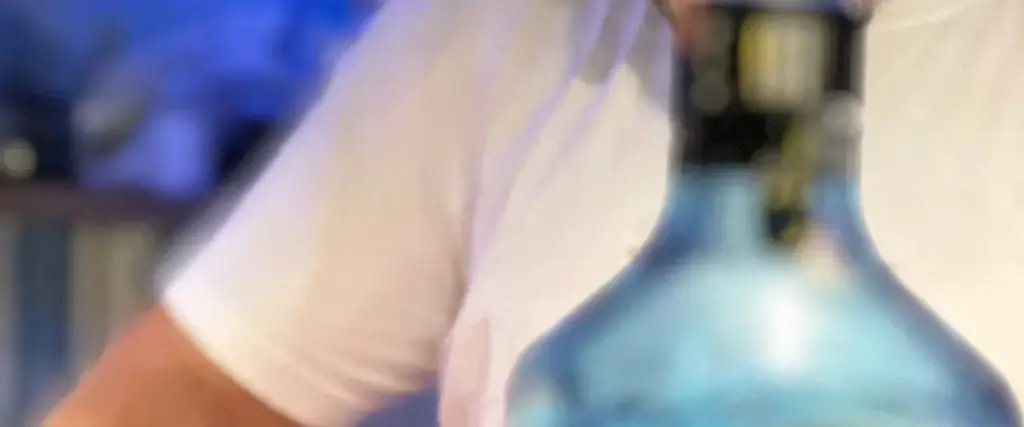 A bartender in a white T-shirt at the counter, a pale blue glass bottle standing in front of him, the room behind out of focus