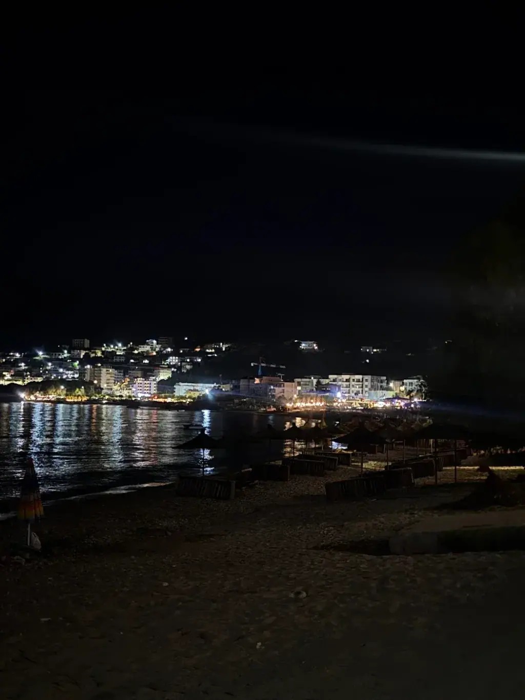 Potam beach at night with folded parasols on the pebbles and the lights of Himarë across the water