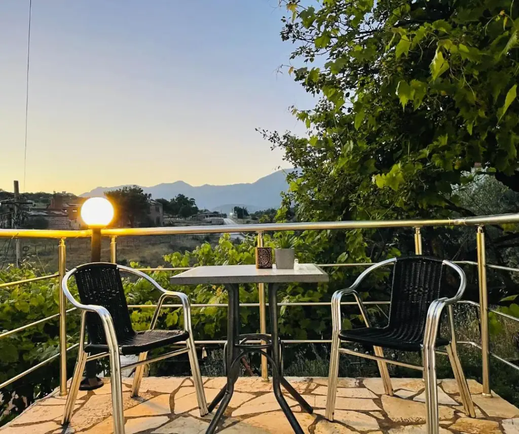 Two black bistro chairs and a table on the hotel's flagged terrace at dusk, a globe lamp lit on the balcony rail and the mountains behind
