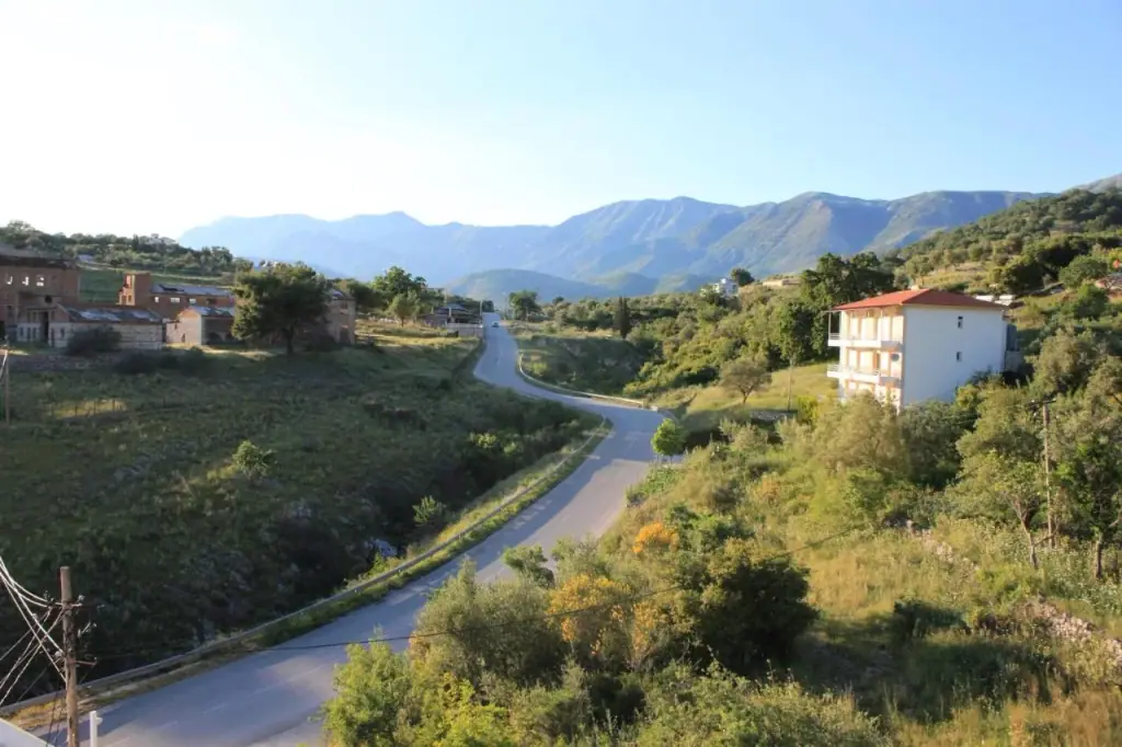 The SH8 curving down between green slopes, a derelict brick complex with corrugated roofs on the left and the mountain ridge along the horizon