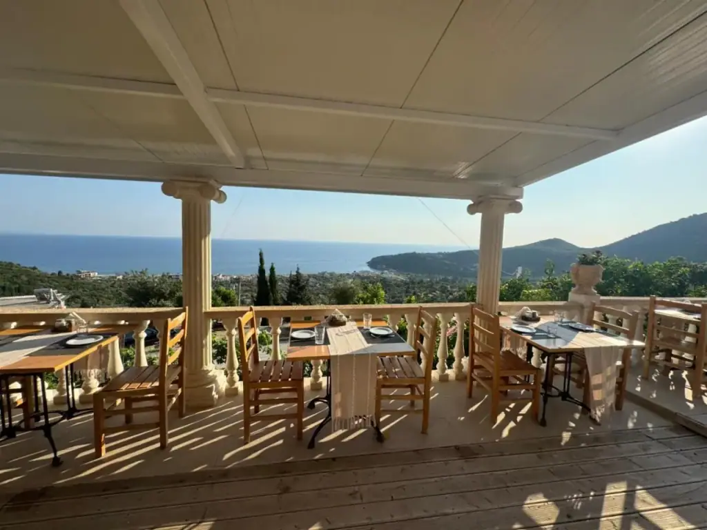 A covered terrace of wooden tables and chairs behind a run of white columns, looking down over the bay