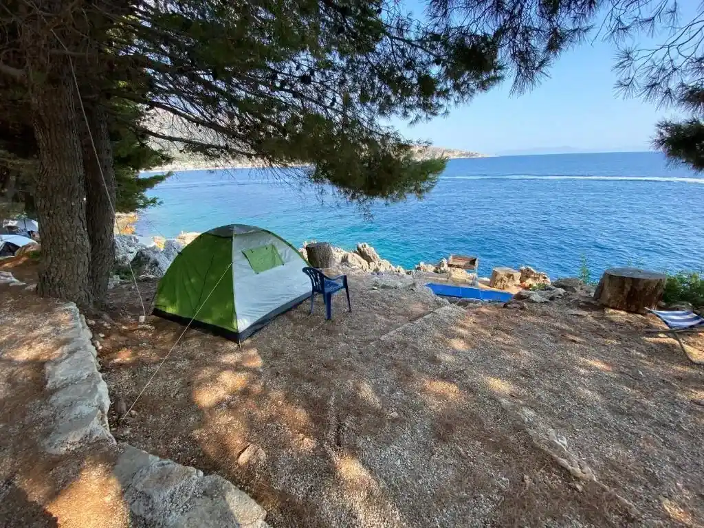 A green and white dome tent pitched on a stone terrace under a pine, sea beyond