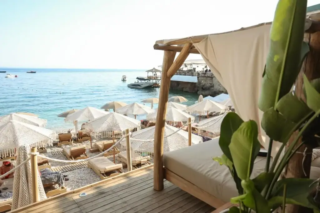 A timber cabana with a canvas canopy and a cushioned daybed on a deck above straw parasols and the sea