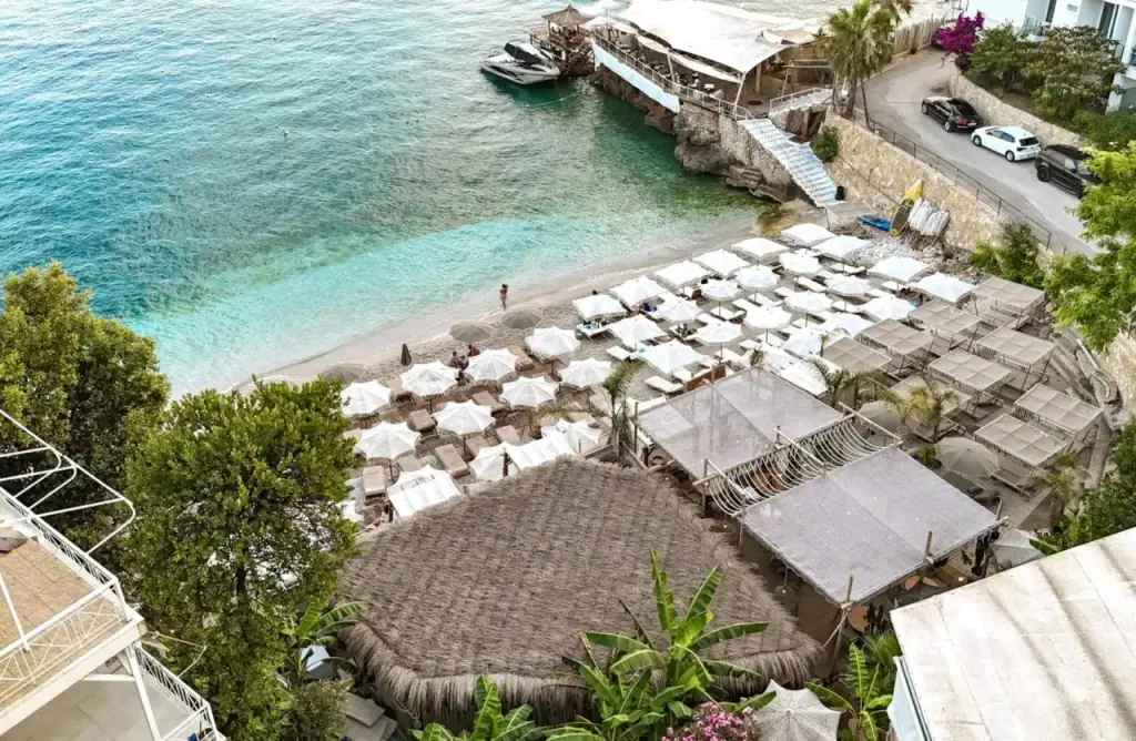 A drone view of a small cove with a boat at a jetty, rows of parasols and a road cut into the slope behind