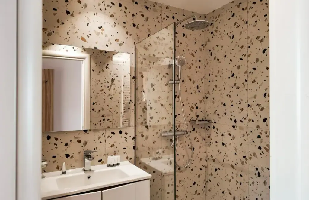 A bathroom lined floor to ceiling in pale terrazzo with dark aggregate, a walk-in shower and a rain head