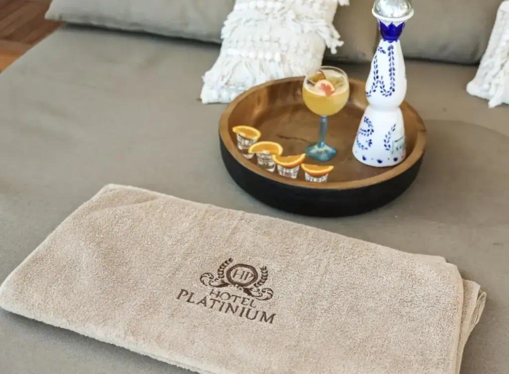 A tray of drinks and a folded towel embroidered Hotel Platinium, on a taupe daybed cushion