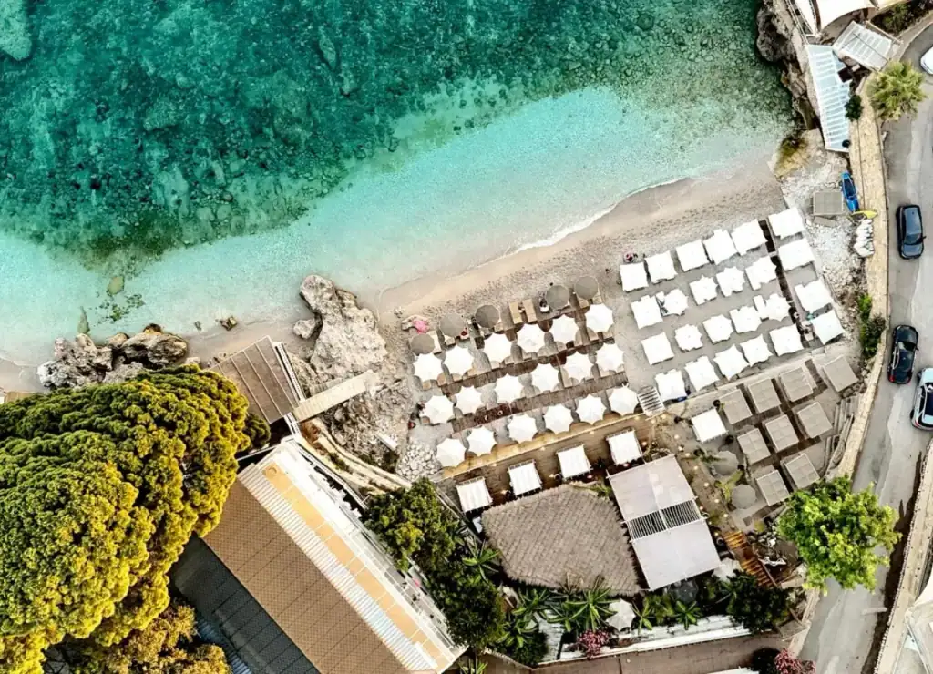 A drone view straight down onto ranks of white square parasols on pale shingle beside clear turquoise water
