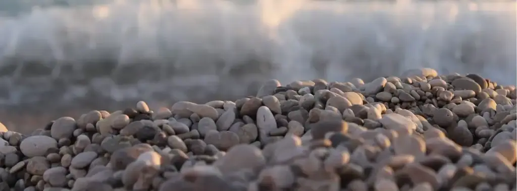 The pebbles of Livadhi beach in close-up, warm tan and grey, the surf breaking behind them