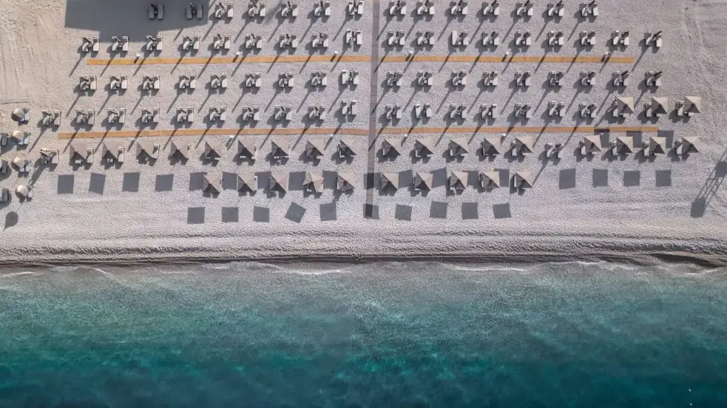 The hotel's beach from directly overhead, straw umbrellas in ruled rows on pale shingle with the shoreline running across the frame