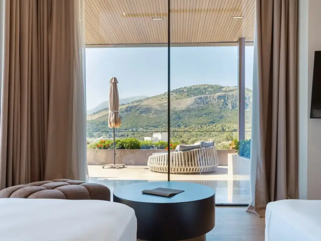 The oversized furnished balcony of the Deluxe Suite seen from inside, a rattan daybed and a folded parasol against the green ridge