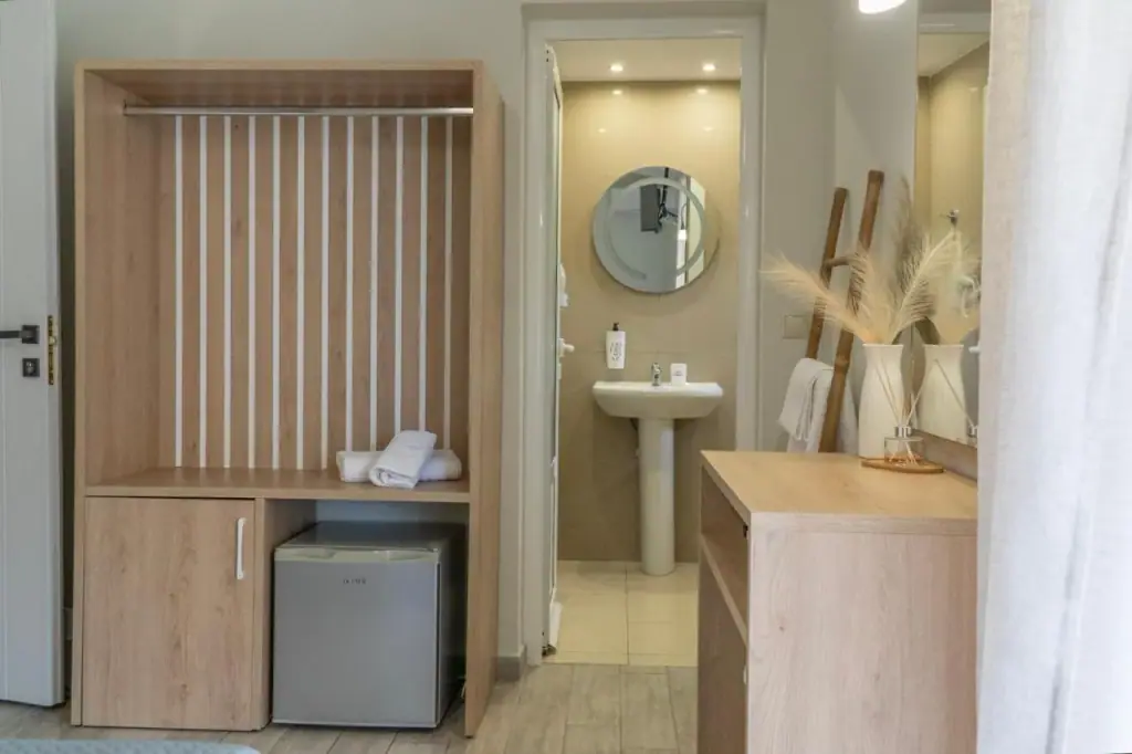 An open oak wardrobe with a fluted back and a small fridge below it, with a tiled bathroom and round mirror through the doorway beyond