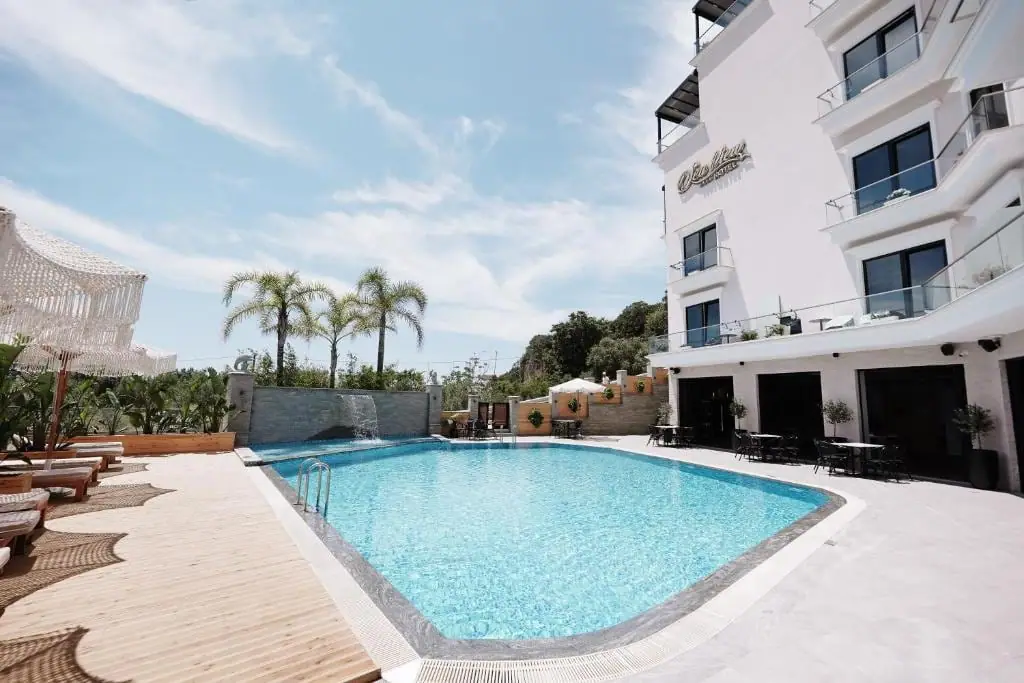 The hotel pool seen along its length, with sun loungers and a fringed parasol on the timber decking to the left, palms behind, and the white hotel and its ground-floor terrace tables on the right
