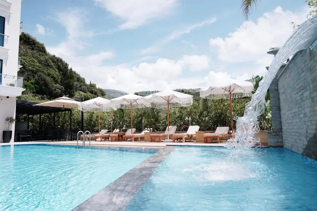 The pool at water level with water arcing from a white dolphin figure set into the stone wall on the right, fringed parasols and timber loungers along the far edge, a black slatted pergola over the bar at the left, and the wooded hillside behind