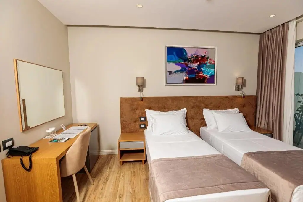 A standard twin room with two single beds under a suede-upholstered headboard panel, an abstract painting above, a sconce either side, and a desk with a framed mirror and a telephone along the near wall