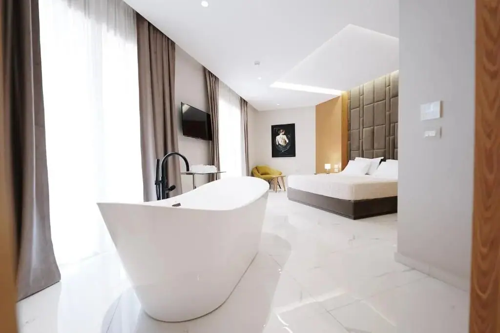 A marble-floored suite with a freestanding white bath in the foreground, a king bed under a quilted headboard beyond, a mustard armchair, a wall-mounted television and full-length drapes