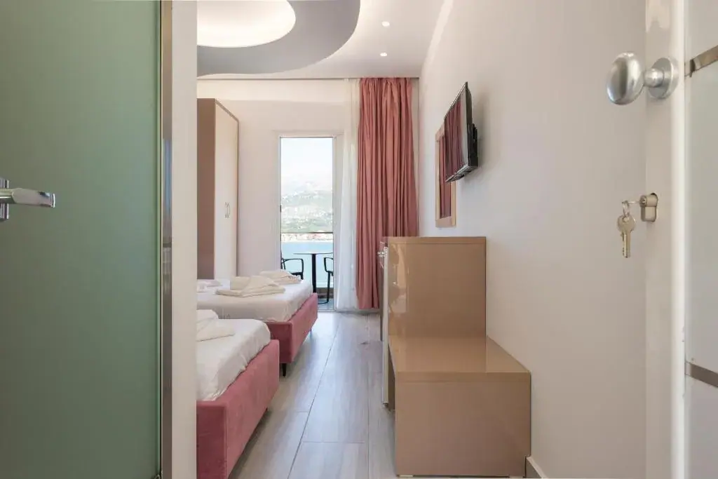 Looking into a room at Seaside Hotel from the corridor, key in the open door, two beds on rose-coloured bases and the bay through the balcony door