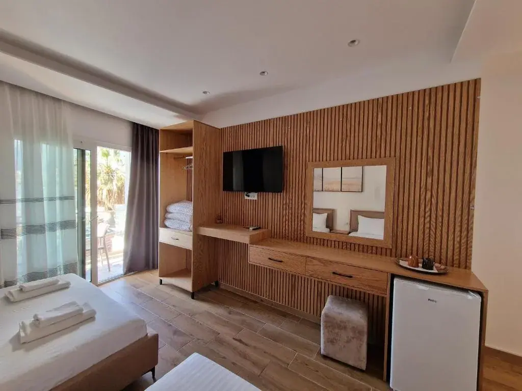 A room at Seaside Hotel whose long wall is oak slatting carrying a television, a mirror, a desk and drawers, with a mini fridge beside it