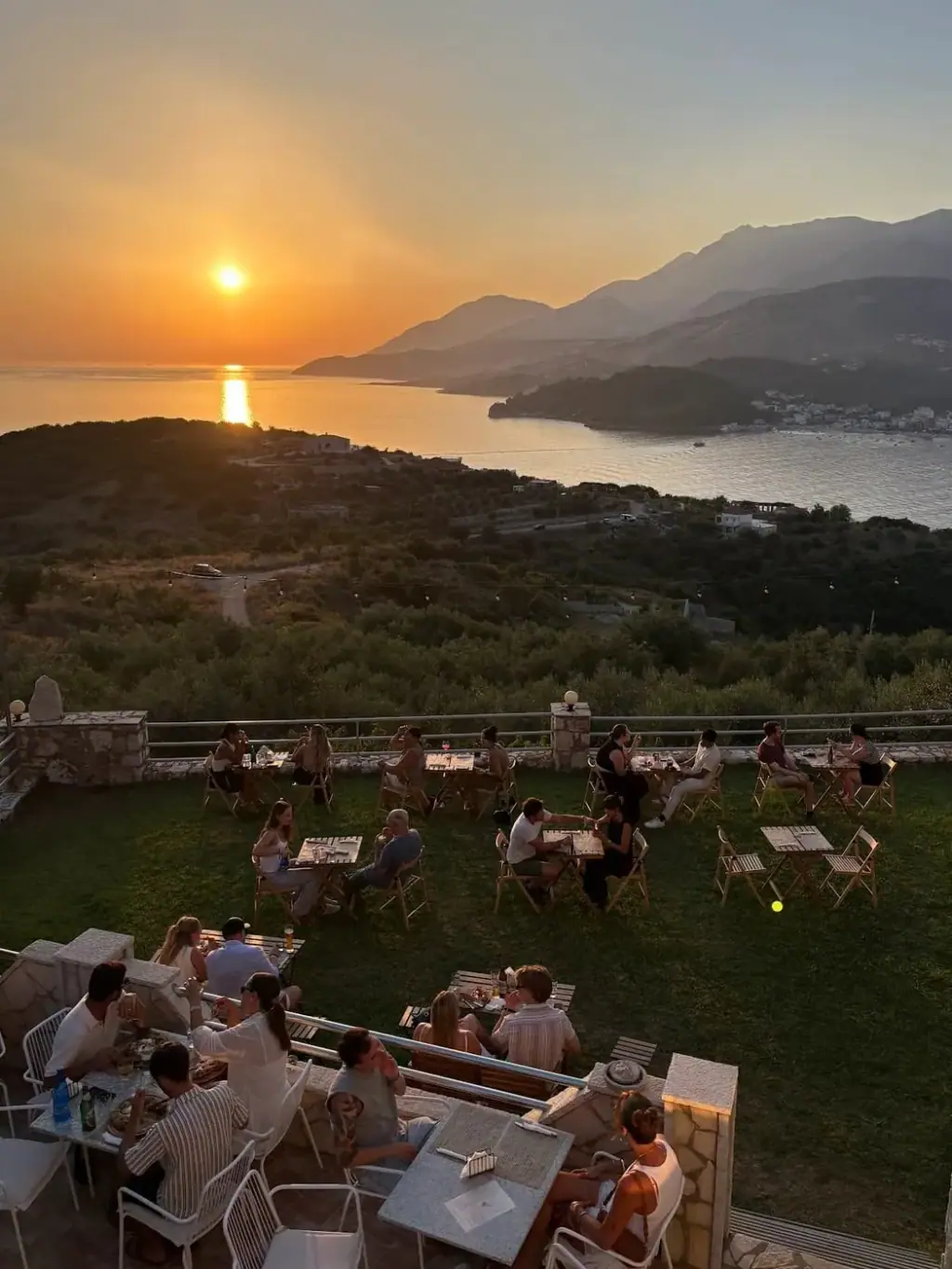 Wooden tables on a grass terrace high above the bay with the sun low over the sea and festoon lights overhead