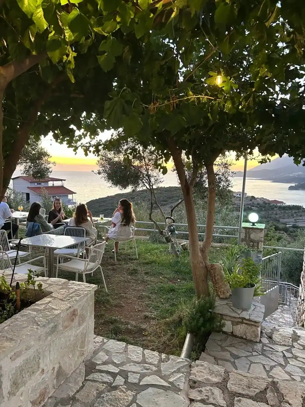 Tables under fig and olive trees on the garden terrace in the evening, the bay behind