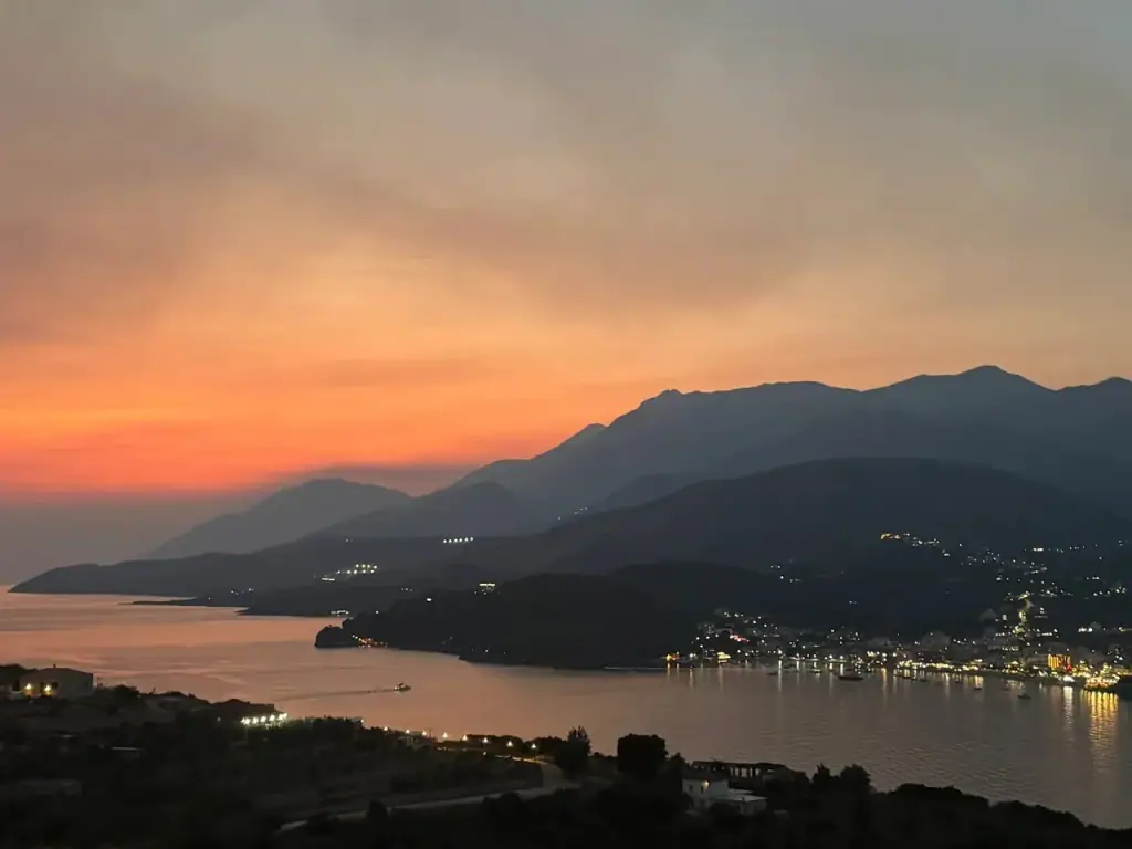 Himarë bay after sunset, orange banding the sky over dark headlands and the town lit along the shore