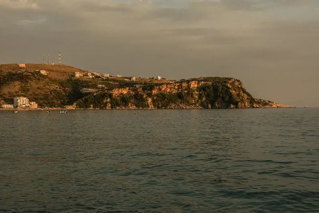 A headland on the Himarë coast in the last of the light, seen across the water, with houses and radio masts along the ridge