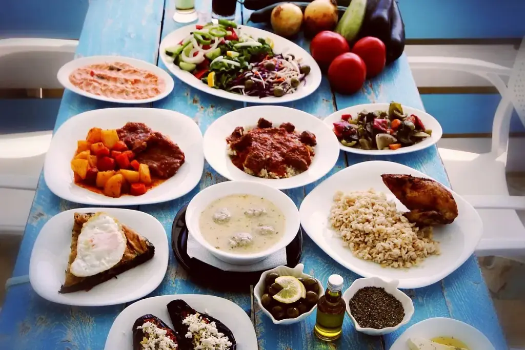 A blue-painted plank table covered with white plates: a chopped salad, a meat stew with potatoes and carrots, meat in tomato sauce over rice, braised mixed vegetables, meatball soup, roast chicken with rice, a wedge of pie under a fried egg, stuffed aubergines, olives, a bottle of olive oil and a bowl of dried herbs