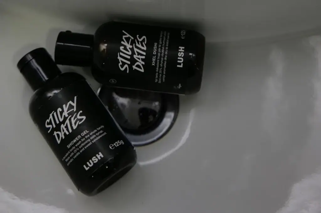Two black Lush Sticky Dates shower gel bottles lying in a white basin, labelled in English and Albanian