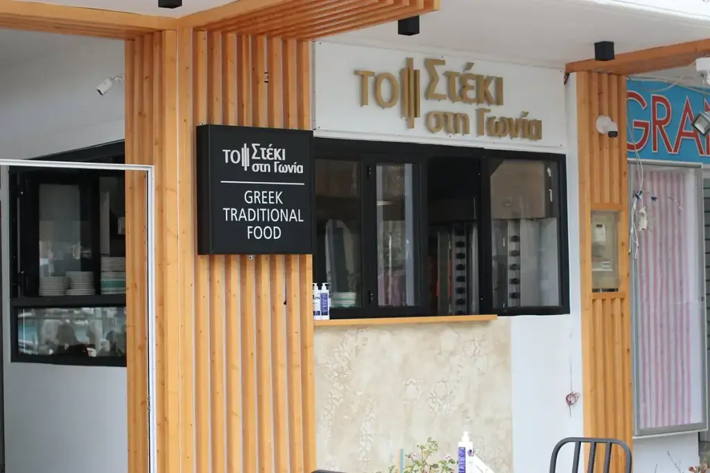 The frontage on Rruga Kaonet: brass Greek letters on white render above a black sign plate, on vertical pine battens