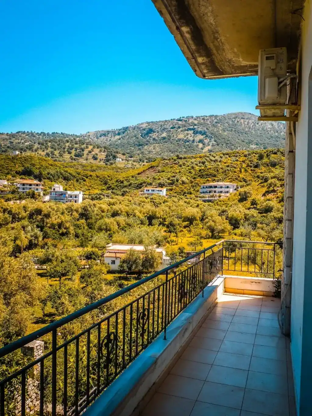 A tiled balcony with an iron rail looking over olive terraces to the mountains behind Himarë