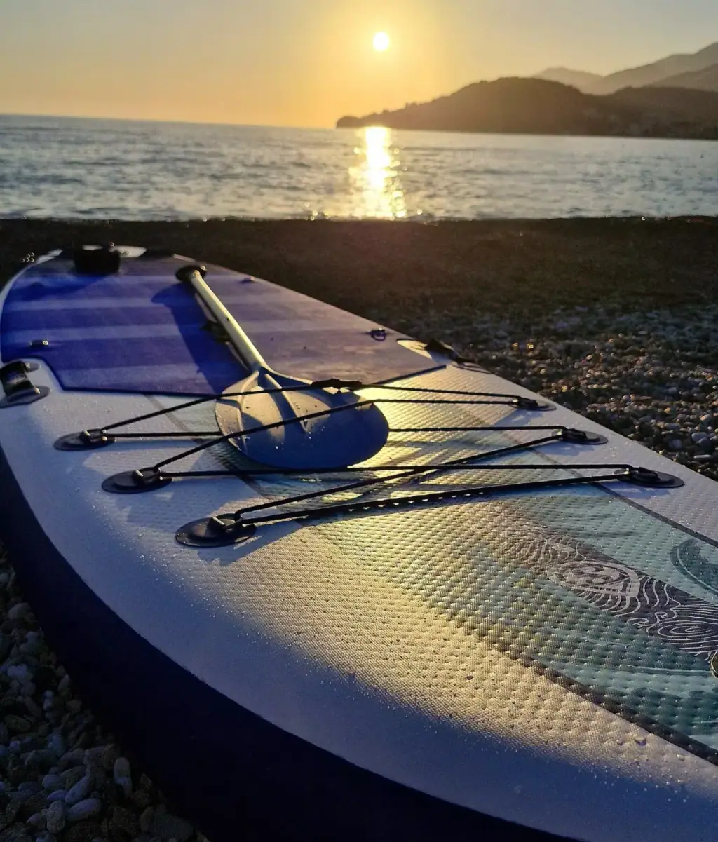A paddleboard and paddle on the pebbles with the sun setting over the Ionian sea