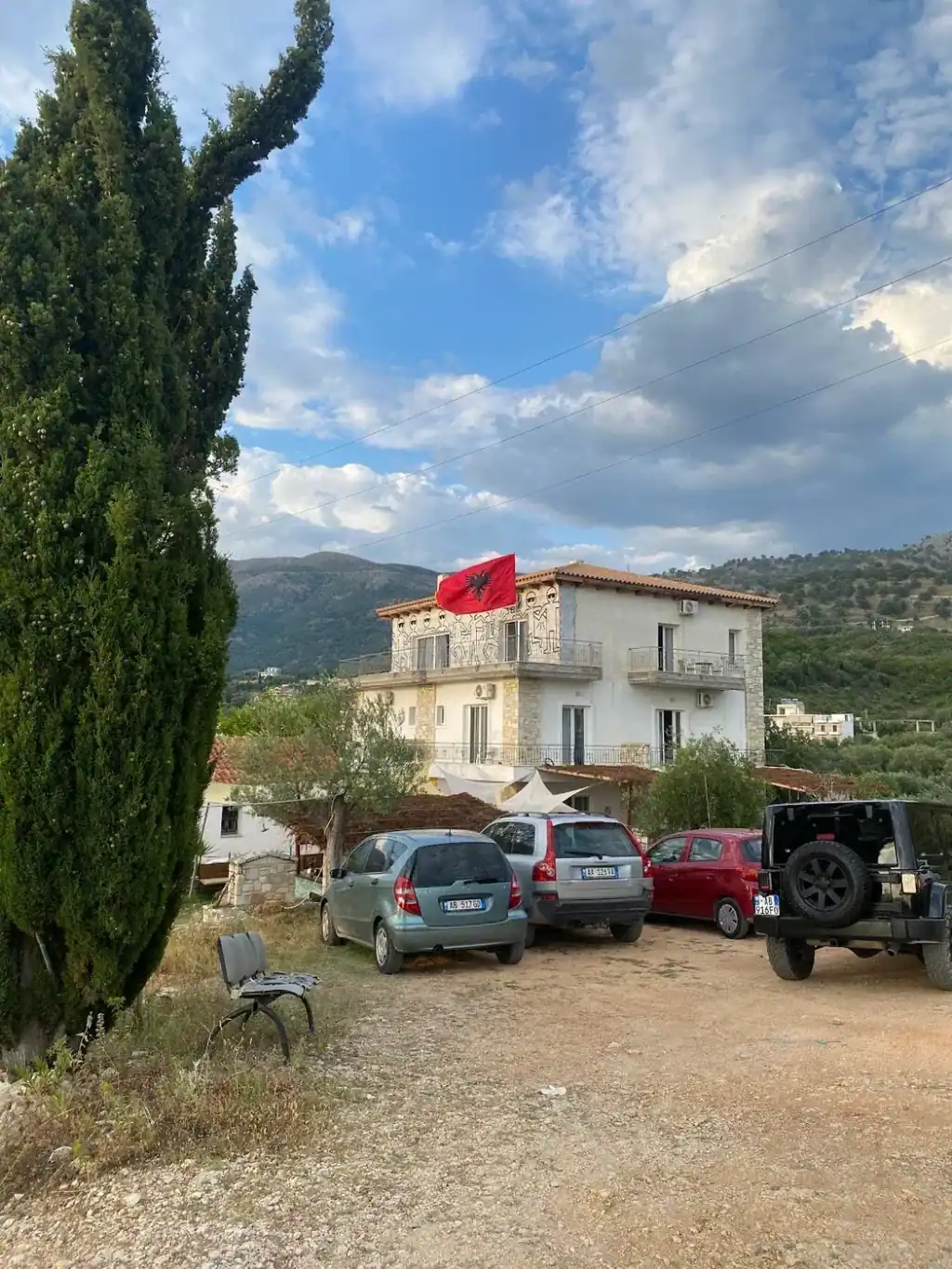 The villa behind a cypress with cars parked on gravel and an Albanian flag on the roof