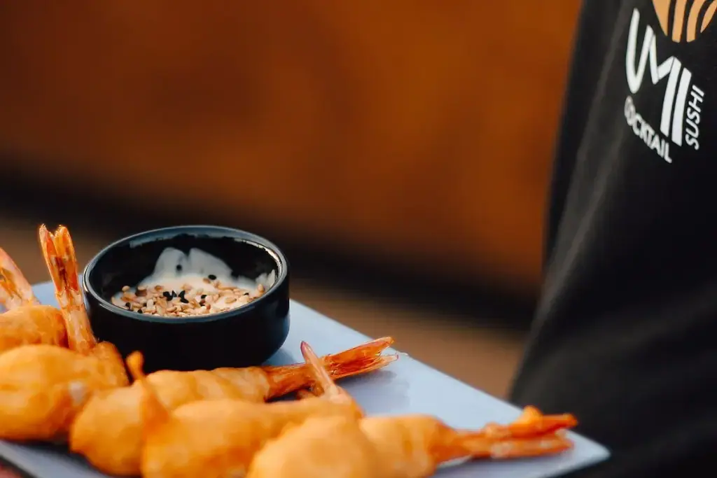 A pale blue-grey plate of prawns fried in batter beside a black bowl of dip strewn with white and black sesame, a black apron printed with UMI's gold fish mark standing behind it