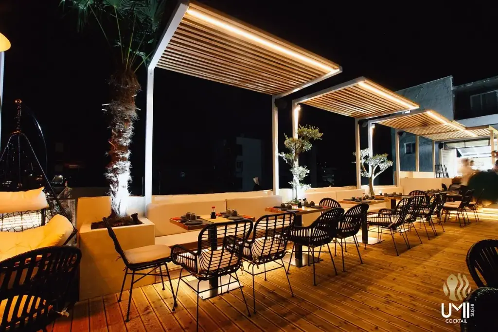 The deck after dark, three timber pergolas lit along their undersides, black rattan chairs at set tables and olive trees in the planters
