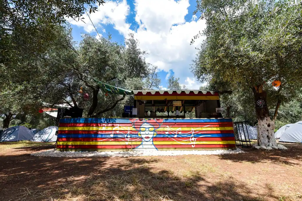 The camp's counter painted in blue, red and yellow planks with a mural of a woman in yellow sunglasses and music notes, under olive trees with white tents behind