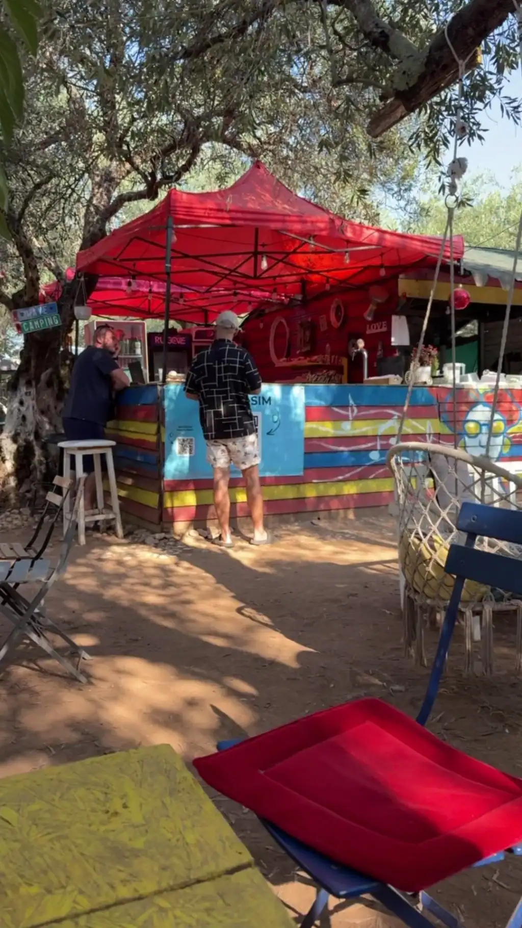 Two guests at a striped counter under a red gazebo in the olive grove, with folding chairs in the shade