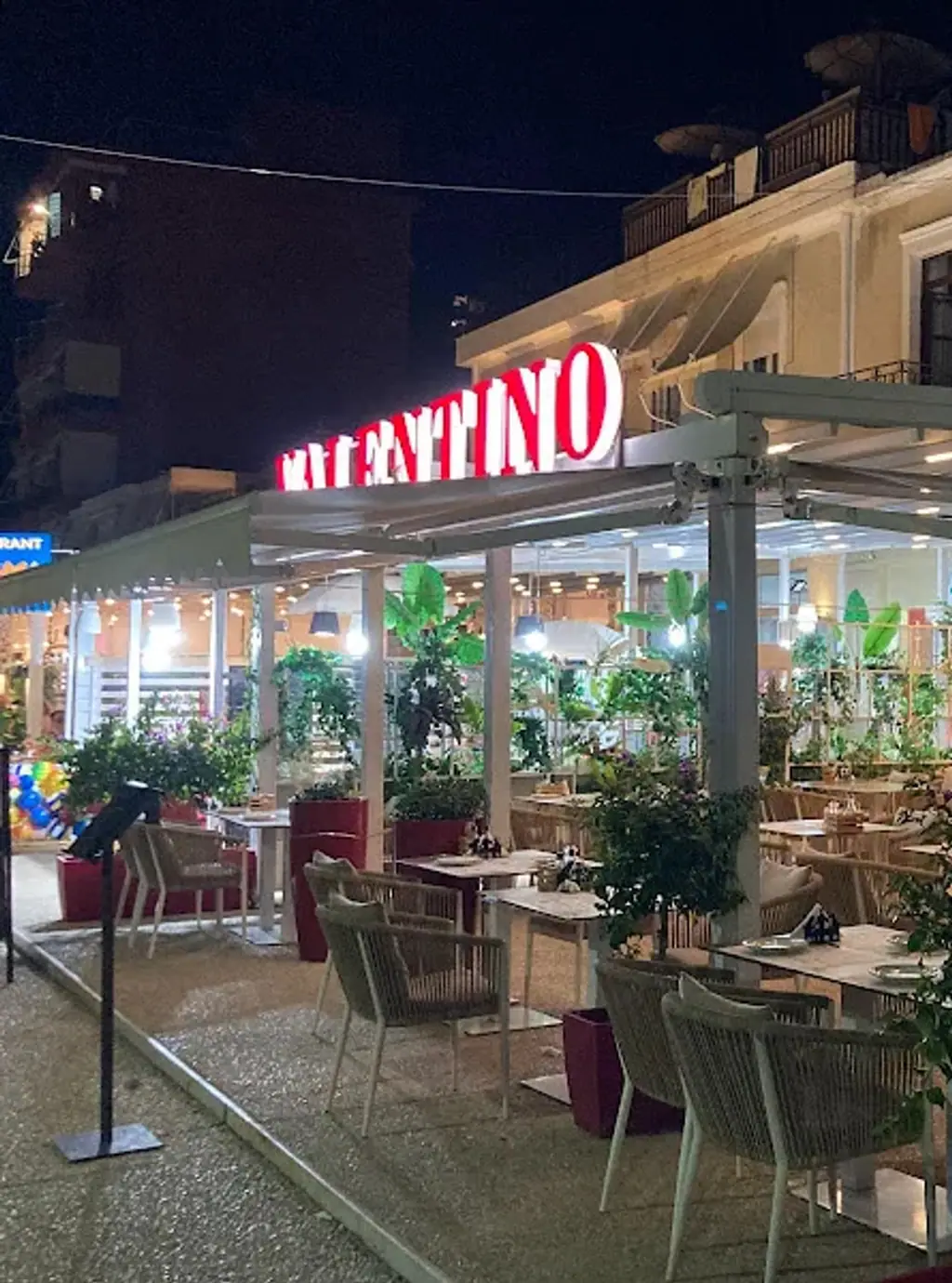 The VALENTINO sign lit above the pergola on the Himarë seafront at night, red letters with white-lit faces, set tables and rope-weave armchairs beneath