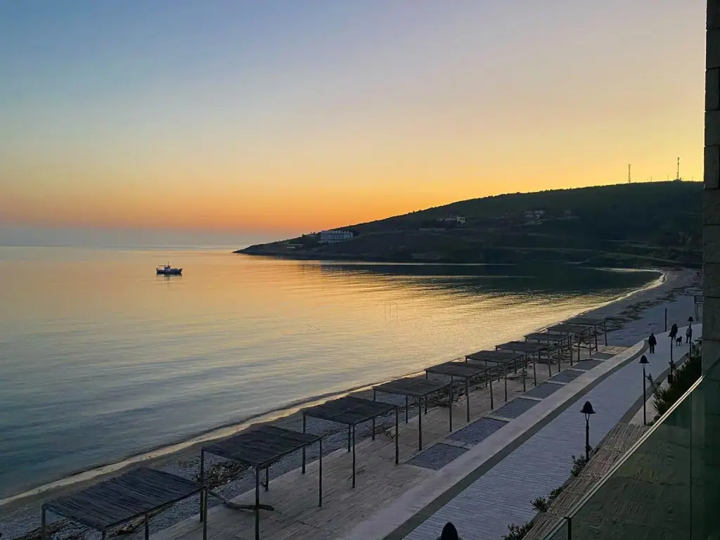 The Qeparo shore after sunset, the promenade lamps still unlit and the flat bay holding the last of the orange sky