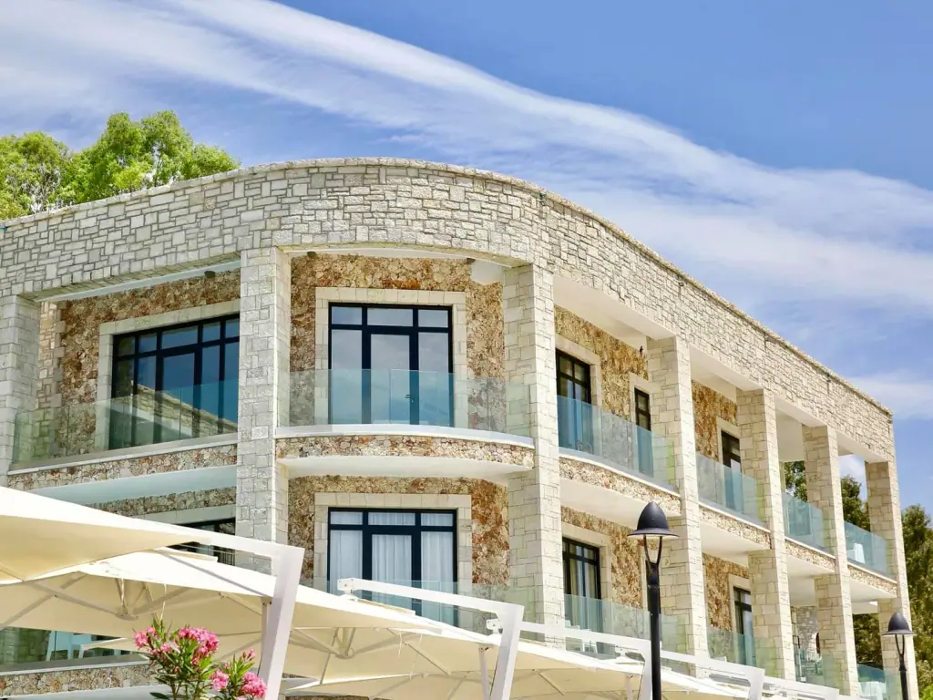 The curved stone-clad corner of the newer front, its square black-framed windows set behind glass balustrades, with white fabric parasols on the terrace below