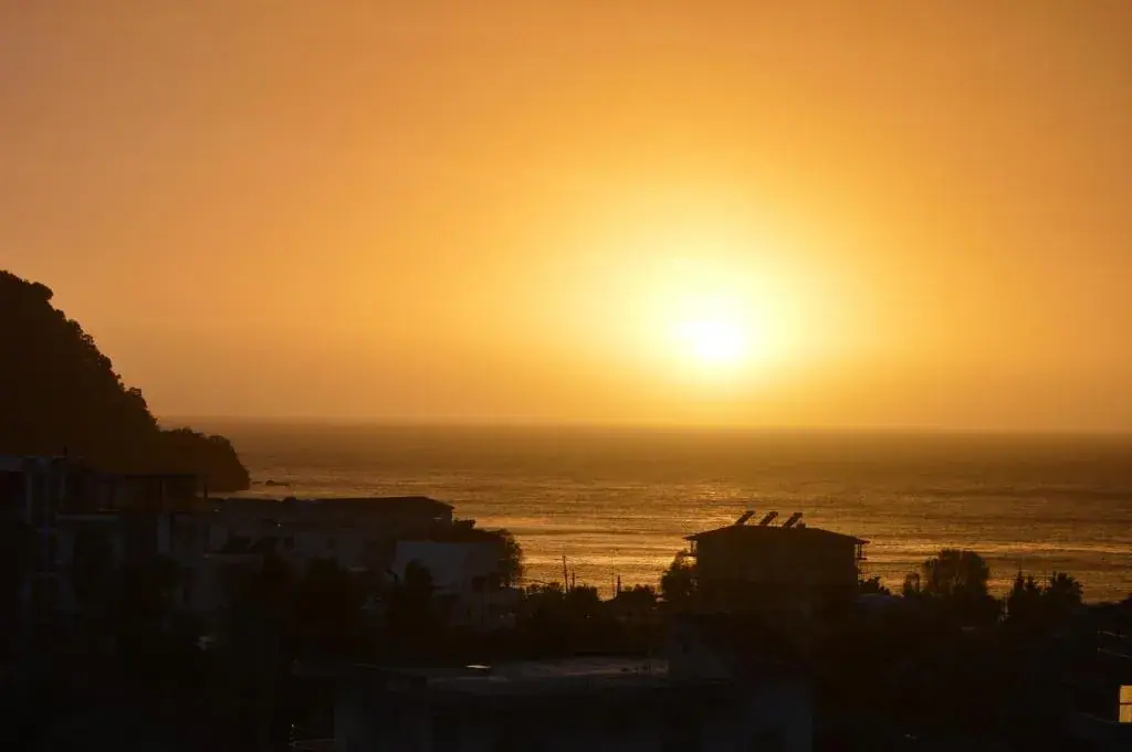 The sun setting into the Ionian off Potam, low rooftops and a wooded headland in silhouette below
