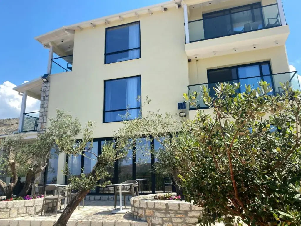 The cream-rendered building from the garden, two storeys of glass-balustraded balconies above a glazed ground floor, olive trees in front